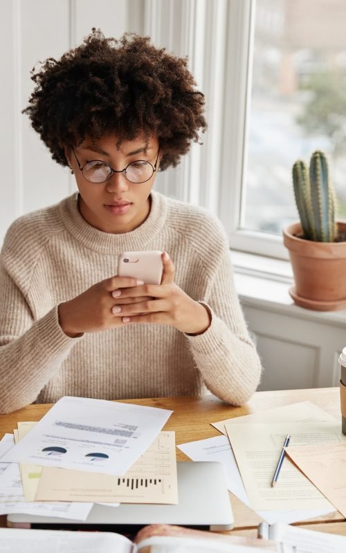 busy-businesswoman-with-afro-hairstyle-works-with-documentation-checks-bank-account-via-cell-phone.jpg